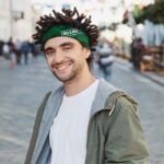 Young handsome bearded man dressed stylish casual clothes, white t-shirt, green jacket, bandana posing outdoor. Smiling hipster with dreadlocks hairstyle, close up portrait, street photo, urban style